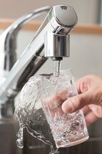 Das Bild zeigt einen laufenden Wasserhahn aus chrom. Die rechte Hand von einer Person hlt unter diesen Wasserhahn ein Glas und das Wasser schwappt sprudelnd ber.