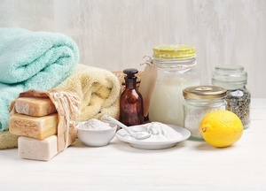 Das Gesamtbild zeigt Handtcher und Reinigungsprodukte bzw. Dinge, die zu deren Herstellung ntig sind. Auf der linken Seite sieht man im Hintergrund ein mintgrnes und ein gelbes Handtuch. Davor liegen 3 feste Seifenstcke, lose in ein braunes Papier eingepackt. In der Bildmitte befindet sich eine kleine braune Flasche mit Stpsel. Davor steht eine weie Keramikschssel und ein weier Keramikteller mit einem Lffel. Beide Keramikgefe enthalten ein weies Pulver. Rechts befinden sich 3 Glser in unterschiedlichen Gren und Formen. Das eine Glas beinhaltet eine Seifenflssigkeit, das andere Kruter. Vor dem dritten Glas liegt eine Zitrone. Der Inhalt ist hier nicht ersichtlich.AdobeStock_282569640