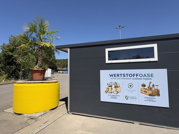 Das Bild zeigt im rechten Teil das Geb�ude der Wertstoffoase. Das Geb�ude ist antrhazitfarben hat ein wei�es Oberlicht und ein Flachdach und im unteren Teil befindet sich das Schild mit dem Titel "Wertstoffoase - Gutes weitergeben, Sch�nes finden. Abgebildet sind auf dem Schild in Holzkisten auch Gegenst�nde wie Geschirr, ein Skateboard, ein Fu�ball, eine Gitarre und ein Roller. Links dem Geb�ude befindet sich ein Betonsockel, der signalgelb angestrichen ist und auf dem eine gro�e F�cherpalme steht. Im Hintergrund sind Teile des Recyclinghofs K�ssaberg zu sehen.