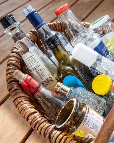 Auf dem Bild ist ein Korb zu sehen der auf einem Holzfußboden steht. Im Korb liegen Verpackungsgläser verschiedener Farben und Formen. altglas-marcella-merk-initiative-muelltr-wirkt