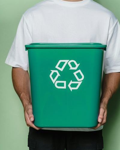 Auf dem Bild ist der Oberkörper einer männlichen Person mit weißem T-Shirt und schwarzer Hose zu sehen, die eine grüne Box mit dem Recycling-Symbol in den Händen hält. pexels-lara-jameson-9324359