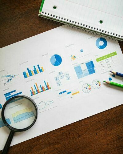 Das symbolische Bild zeigt verschiedene Diagramme&comma; eine Lupe und zwei Buntstifte in blaund gr&uuml;n sowie in der rechten oberen Ecke einen Spiralblock