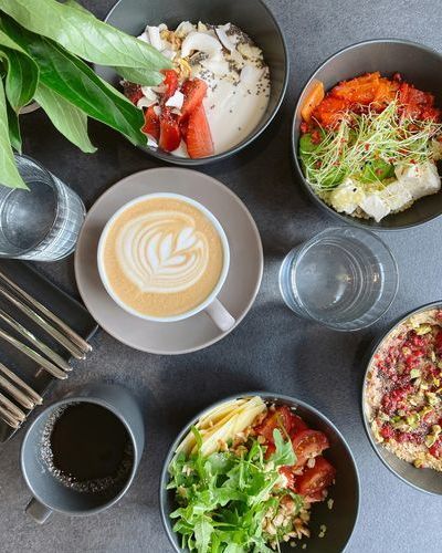 Auf dem Bild ist ein grauer Tisch mit verschiedenen grauen Schüsseln mit Lebensmitteln zu sehen, sowie eine Capuccino-Tasse, eine graue Tasse mit Kaffee und zwei Gläser mit Wasser. pexels-valeriya-kobzar-42371713-8257439