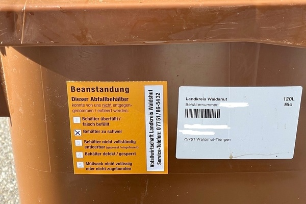 Gezeigt wird ein Teilausschnitt einer braunen Biotonne, die mit einem orangefarbenen Beanstandungsaufkleber versehen ist, auf dem "zu schwer" angekreuzt ist.